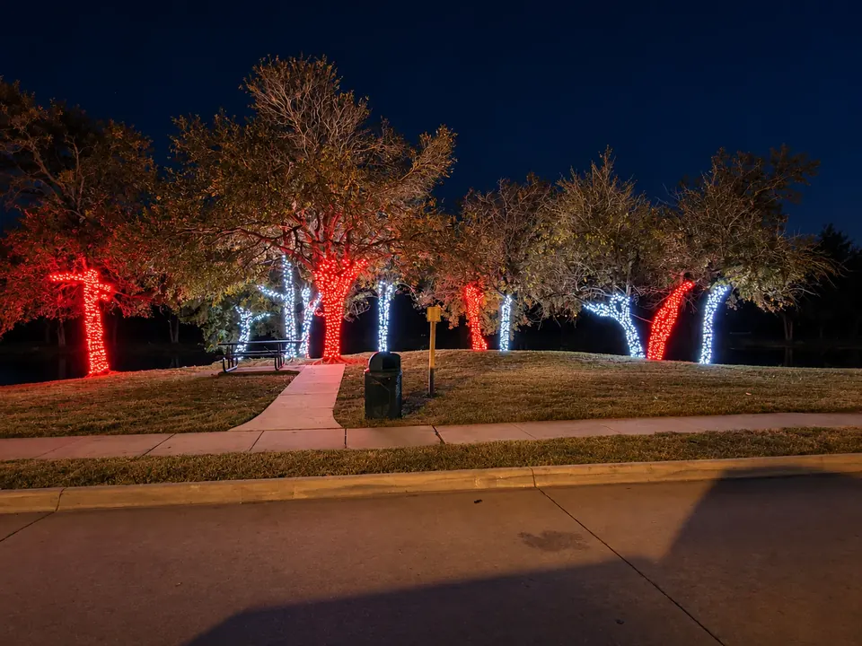 Trees wrapped in red and white Christmas lights beside a community park path